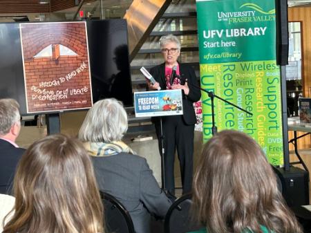 Dr. Joanne MacLean stands in front of an audience. She has grey hair and is wearing a Pink Anti-Bullying Shirt under a black suit. She is holding a book and talking about Freedom to Read
