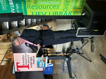 A man is wearing a pink anti-bullying shirt under a black suit. He stands at a podium, holding a banned book and is reading from it.