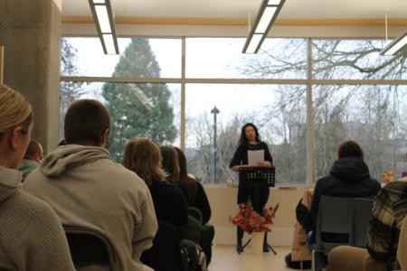 Author Leanne Dunic stands in front of an audience. She had dark hair and is wearing a dark outfit. She holds one of her books as she discusses it at the event.