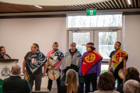 An Indigenous Woman stands at a podium and is partially hidden by the microphone. Five Indigenous men in different regalia stand to her left.