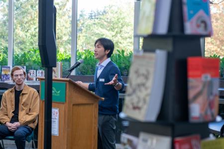 Japanes man with dark hair and a blue cardigan over a blue dress shirt stands at a wooden podium and speaks into a microphone. In the foreground, a display of Japanese books is out of focus but are part of a donation of books from the Consulate General of Japan in Vancouver.