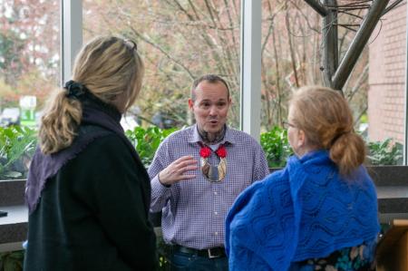 In the foreground, the backs of two women are towards the camera. One woman has blone hair tied back into a ponytail and is wearing a black cardigan. The other woman has auburn hair also pulled back and is wearing a blue scarf as a sweater. Facing the camera, between the woman is a man wiht very short brown hair. He is talking and you can see dark tattoos on his neck.