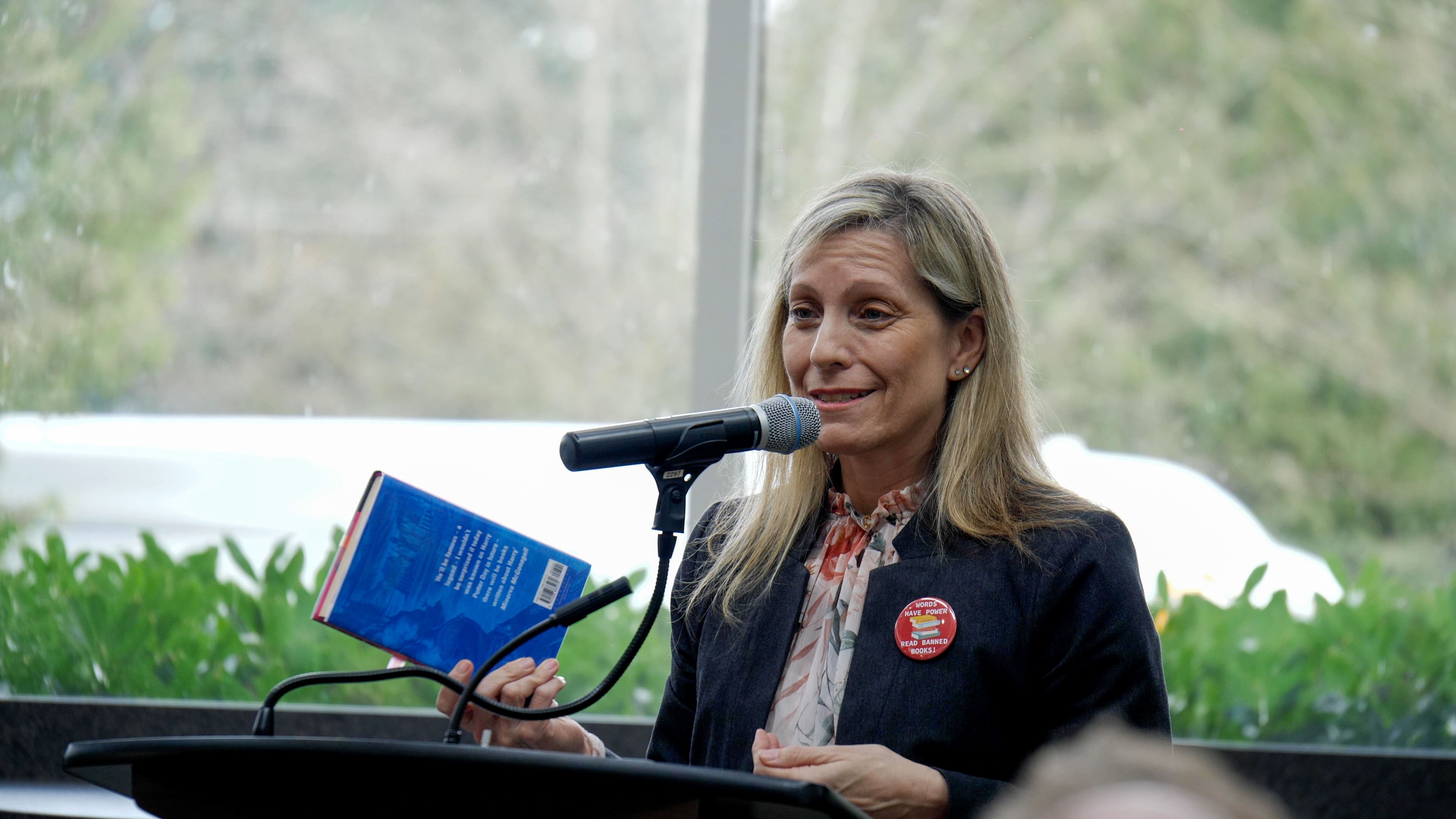 A blond woman stands with a microphone in front of her mouth. She's speaking at a Freedom to read event. A blue book is held in her right hand in preparation of being read out.