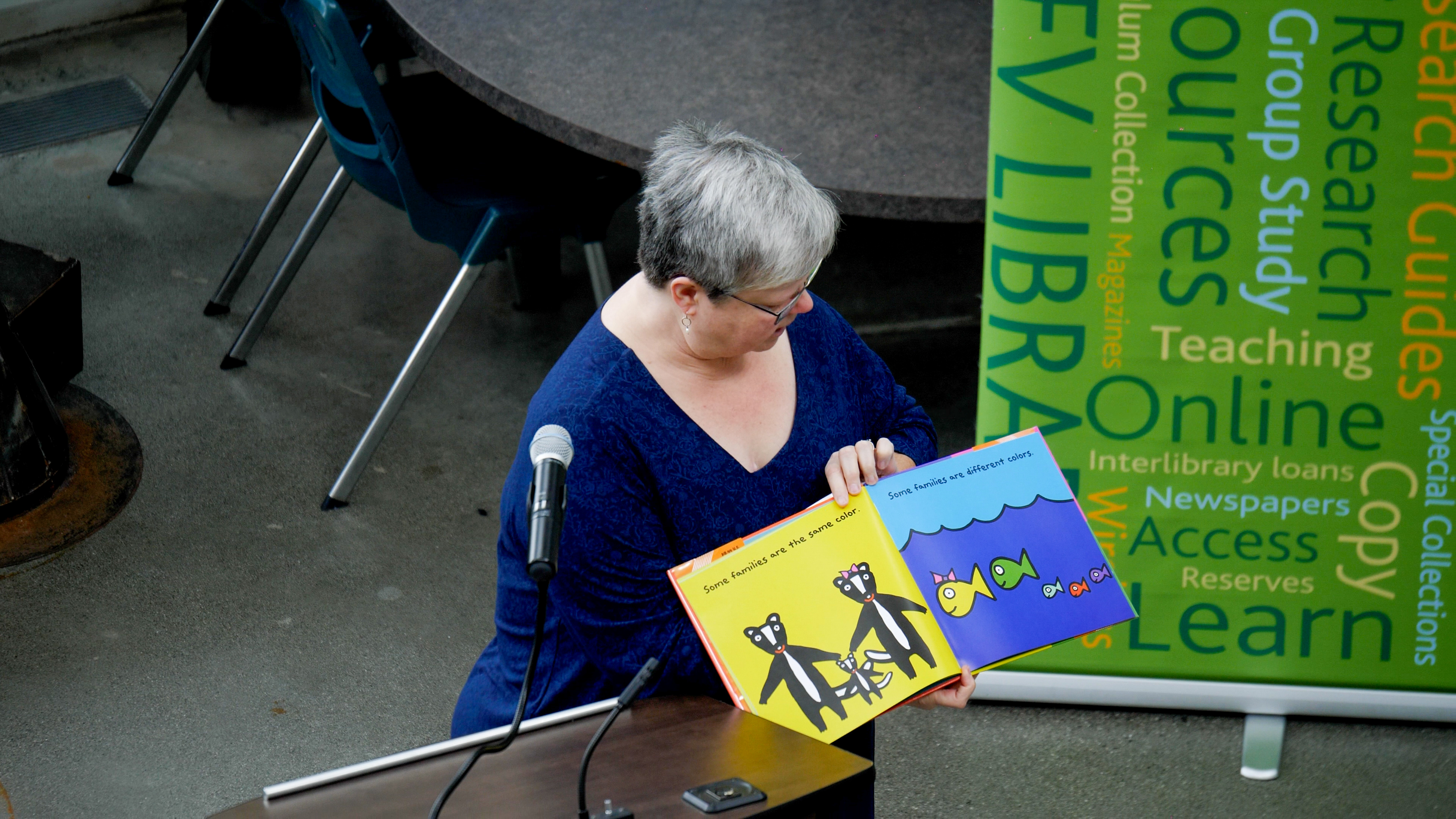 Grey and white haired librarian is wearing a blue shirt. She holds up a colourful book as she reads it out at a Freedom to Read event.