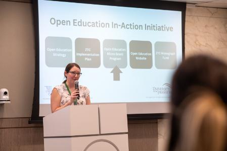 A brown haired woman stands at a white podium. She holds a microphone and is smiling. Behind her is a slide that reads Open Education Iniatives