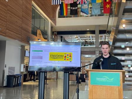 A male student wearing a SUS jacket stands at a wooden UFV podium. He is speaking into a mic. Besides him a digital display shows a freedom to read poster.
