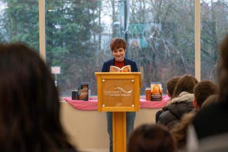 A young woman with light brown hair, wearing a red shirt with a blue dress jacket stands at a wooden UFV podium. She holds an open book and is smiling at the audience. The backs of several heads of students and faculty face the camera.